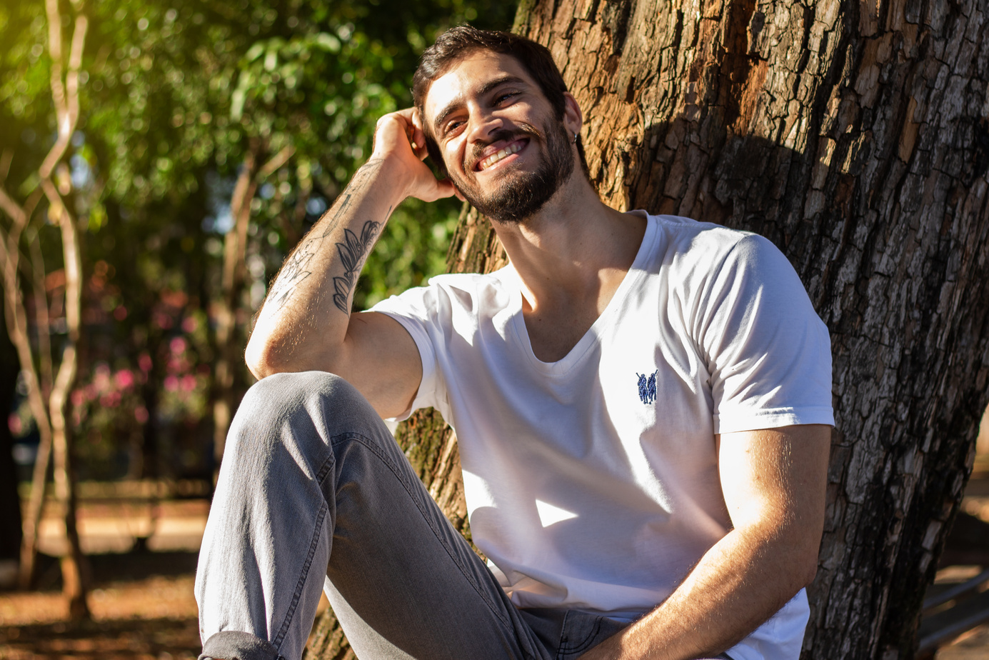 Stylish tattooed happy man resting near tree trunk